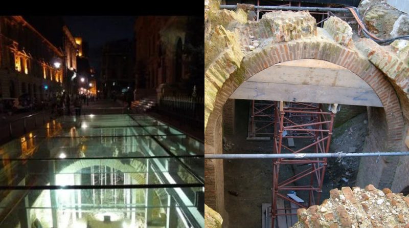 Vestigii arheologice sub sticlă pe o stradă iluminată noaptea și arc roman restaurat cu schele de susținere