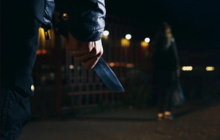 Persoană cu cuțit în mână pe o stradă întunecată, cu o altă persoană în fundal