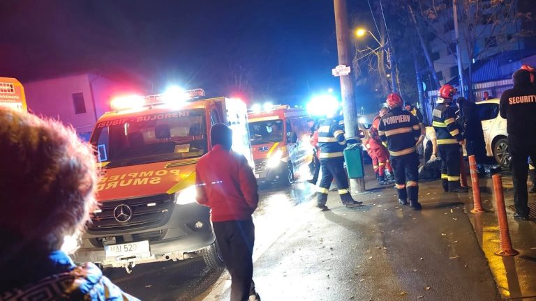 Echipaje de pompieri și salvare SMURD intervin noaptea pe o stradă din București, asistând o persoană rănită pe trotuar, înconjurați de vehicule de urgență și personal de securitate.