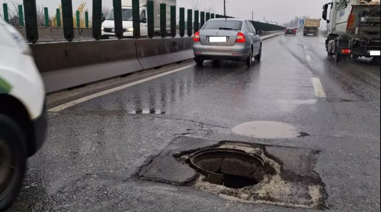 Groapă periculoasă și gură de canal deschisă pe banda de circulație, cu asfalt deteriorat, pe un drum umed, cu trafic auto.