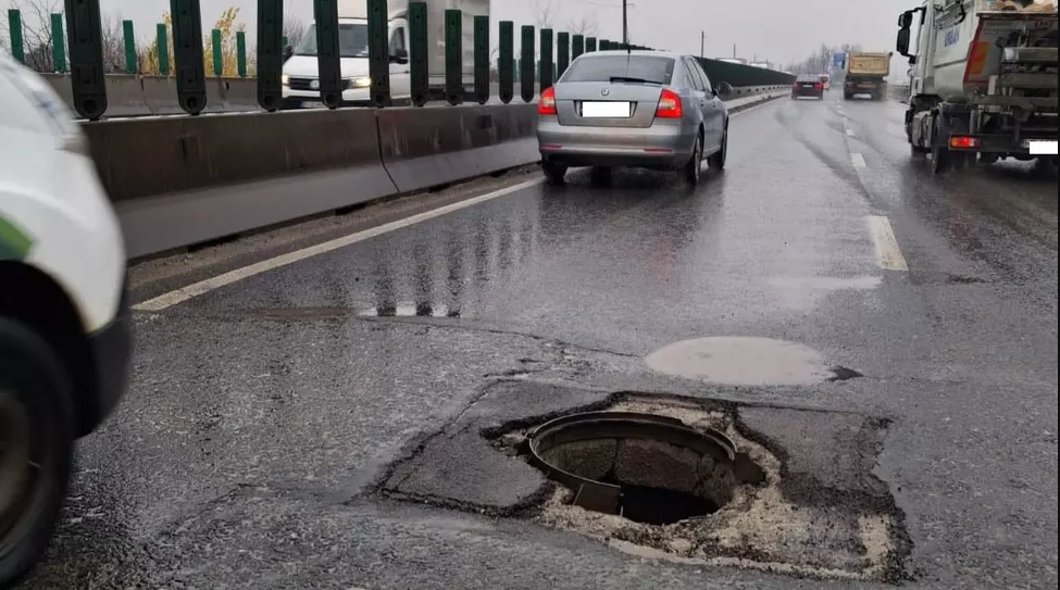 Groapă periculoasă și gură de canal deschisă pe banda de circulație, cu asfalt deteriorat, pe un drum umed, cu trafic auto.