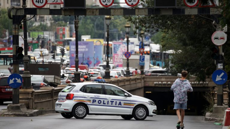 Mașină de poliție (Poliția) din București, România, cu girofarurile stinse, lângă un pasaj subteran aglomerat, în timp ce un copil pe trotinetă trece prin fața ei.