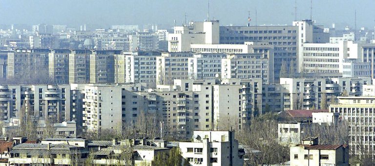 Vedere panoramică a cartierelor de blocuri comuniste din București sub un cer senin, evidențiind densitatea urbană și arhitectura brutalistă specifică perioadei socialiste