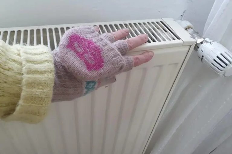 Mână cu mănușă fără degete atinge un calorifer rece lângă un termostat dintr-o cameră.