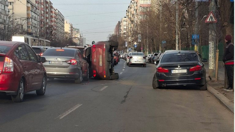 O mașină roșie răsturnată pe o parte în mijlocul unei șosele aglomerate din București, flancată de alte autoturisme și blocuri de locuințe în fundal.