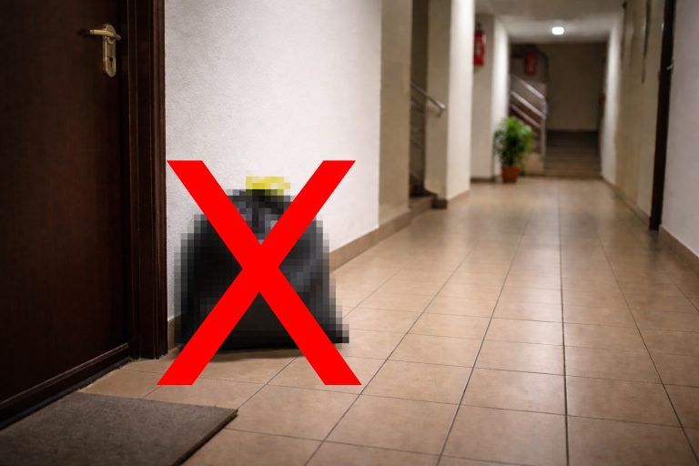 Sac de gunoi negru lăsat pe holul unui bloc lângă ușa apartamentului, marcat cu un simbol X roșu pentru a indica interdicția depozitării deșeurilor în spațiile comune.