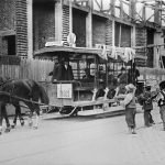 Tramvai hipomobil istoric în București, tras de doi cai, circulând pe lângă o clădire în construcție, cu pasageri și copii pe stradă în perioada interbelică.