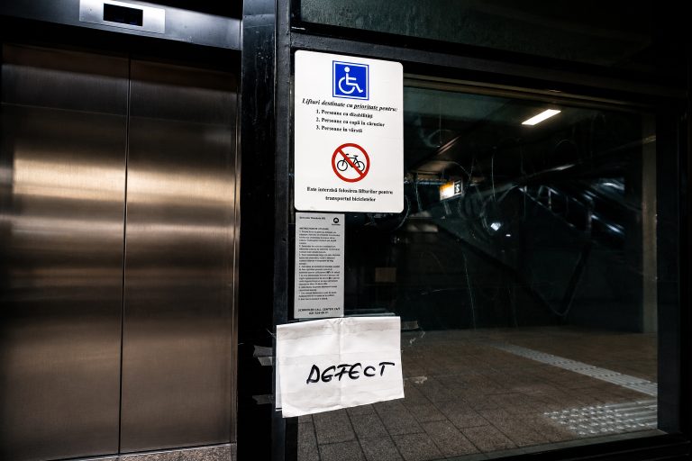 Lift defect în stație de metrou, cu semn pentru acces persoane cu dizabilități și avertizare „DEFECT” lipită pe ușă