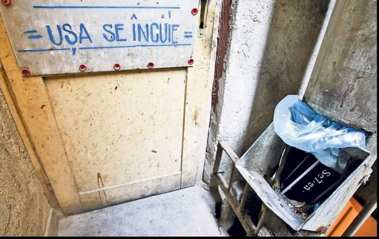 Ușă de acces către ghena de gunoi a unui bloc de locuințe, cu inscripția „Ușa se încuie”, într-o zonă tehnică destinată colectării deșeurilor menajere.