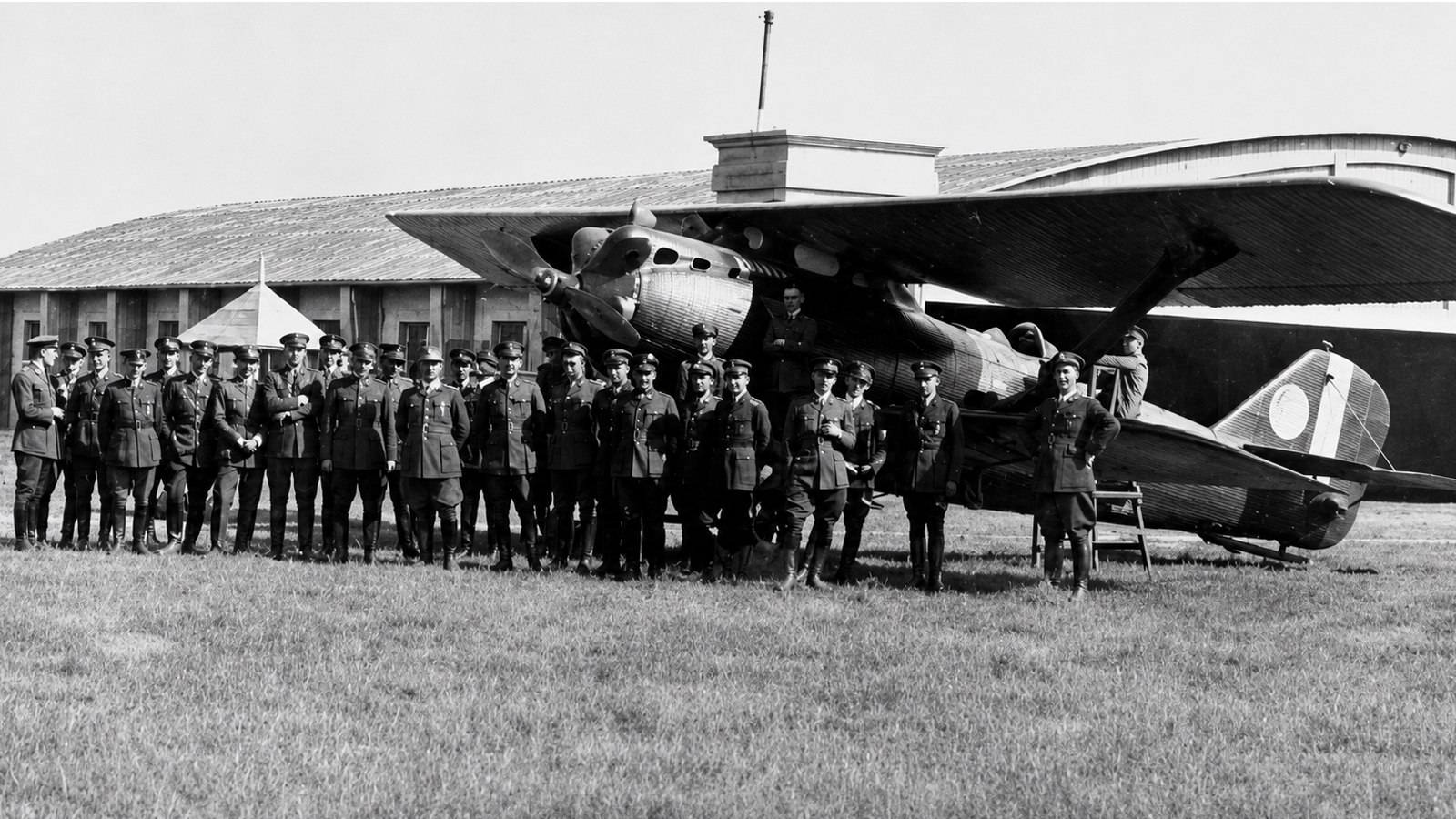 Fotografie istorică alb-negru cu un grup de militari în uniformă pozând în fața unui avion biplan militar pe un aerodrom, în perioada interbelică.