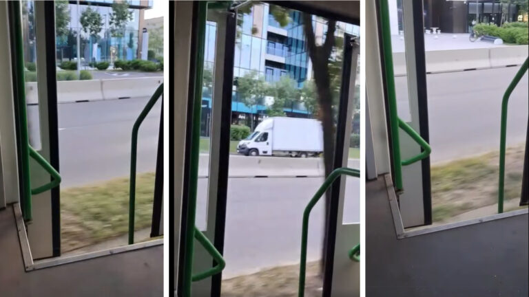 Tramvai în mers cu ușa deschisă, surprins din interior, pe o stradă urbană cu clădiri de birouri în fundal.