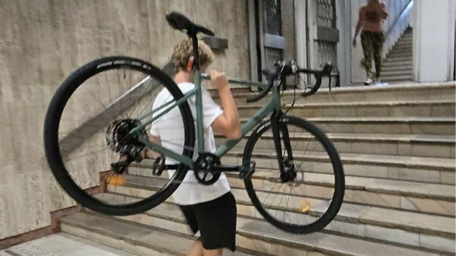 Tânăr care urcă pe scări la metrou purtând pe umăr o bicicletă de culoare verde oliv.